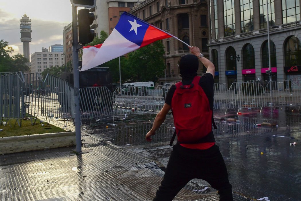 A demonstrator flutters a Chilean national flag during clashes with the police in Santiago, on October 25, 2019, a week after violence protests started. – Demonstrations against a hike in metro ticket prices in Chile’s capital exploded into violence on October 18, unleashing widening protests over living costs and social inequality. (Photo by Martin BERNETTI / AFP)