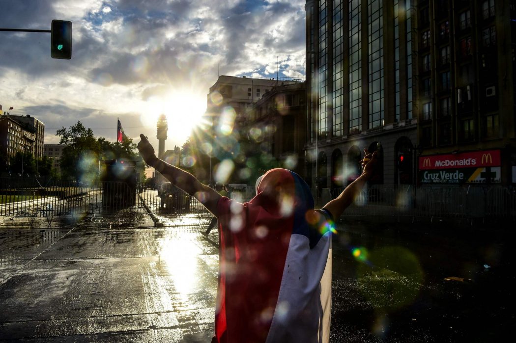 A man wrapped in the Chilean national flag demonstrates in front of police in Santiago, on October 25, 2019, a week after violence protests started. – Demonstrations against a hike in metro ticket prices in Chile’s capital exploded into violence on October 18, unleashing widening protests over living costs and social inequality. (Photo by Martin BERNETTI / AFP)