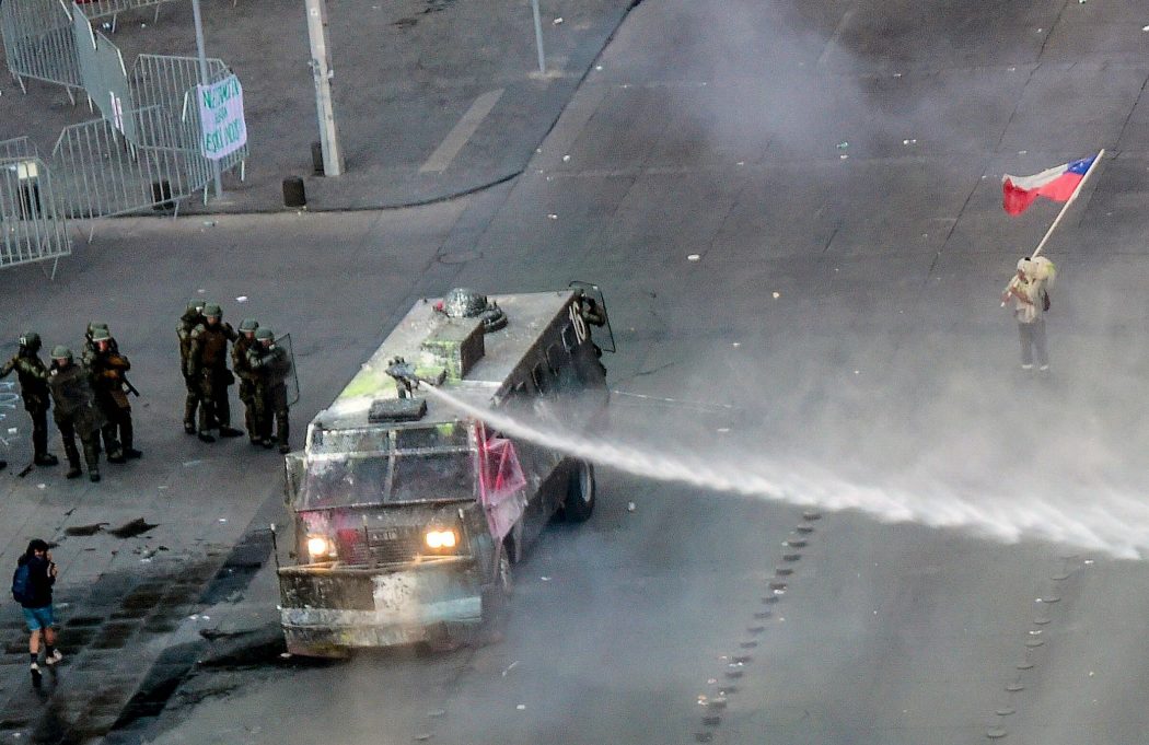 A man demonstrates with a Chilean national flag in front of a police vehicle spraying water in Santiago, on October 25, 2019, a week after violence protests started. – Demonstrations against a hike in metro ticket prices in Chile’s capital exploded into violence on October 18, unleashing widening protests over living costs and social inequality. (Photo by Martin BERNETTI / AFP)