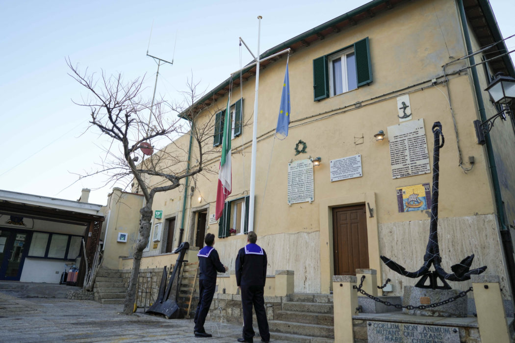 Coast Guard officers raise the Italian flag in the port of the tiny Tuscan island of Isola del Giglio, Italy, Thursday, Jan. 13, 2022. Italy on Thursday is marking the 10th anniversary of the Costa Concordia cruise ship disaster with a daylong commemoration, honoring the 32 people who died but also the extraordinary response by the residents of Giglio who took in the 4,200 passengers and crew from the ship on that rainy Friday night and then lived with the Concordia carcass for another two years before it was hauled away for scrap. (AP Photo/Andrew Medichini)