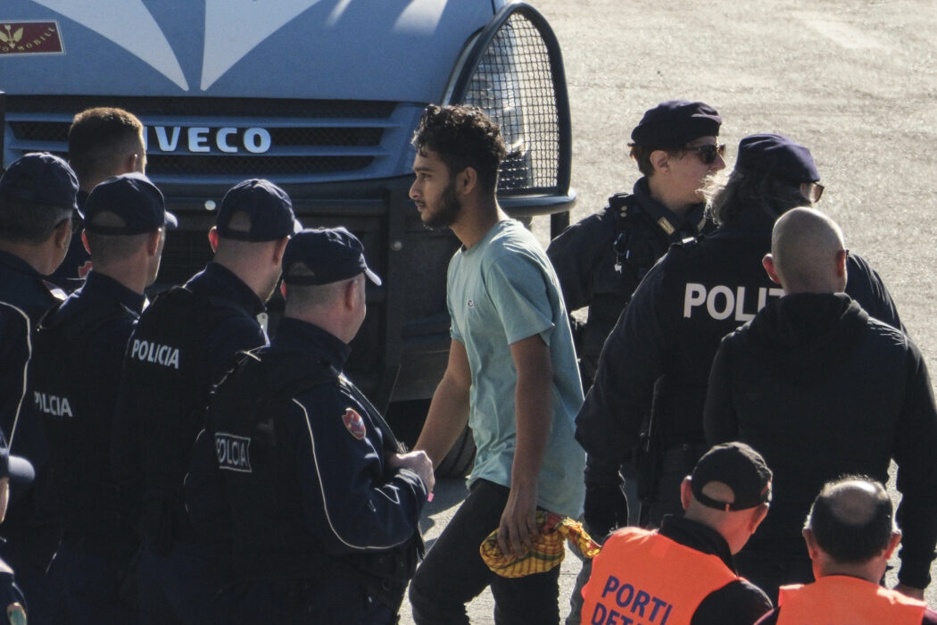 A migrant walks next to security officials at the port of Shengjin, northwestern Albania. Wednesday, Oct. 16, 2024 after he and other migrants disembarked from the Italian navy ship Libra carrying the first group of 16 migrants intercepted in international waters. (AP Photo/Vlasov Sulaj)