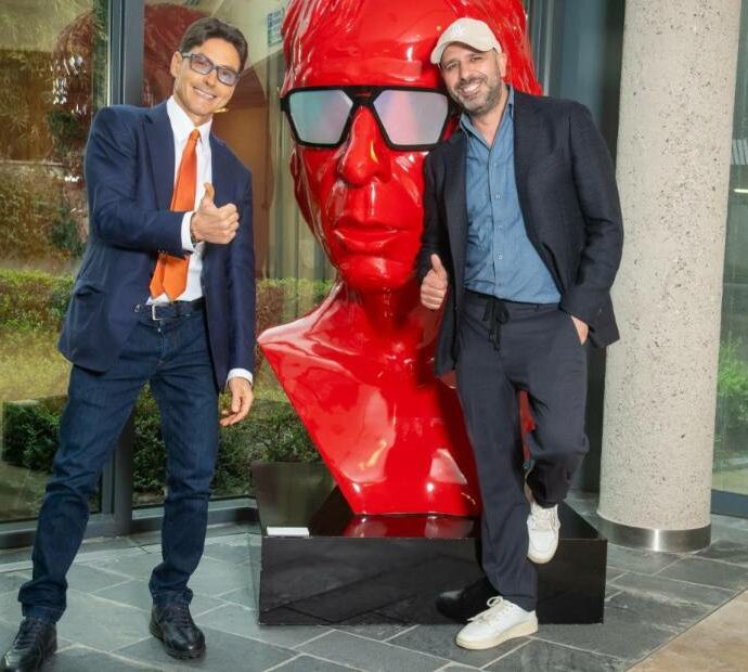 Checco Zalone e Pier Silvio Berlusconi, a sorpresa, si danno il cinque a Cologno Monzese: a “benedirli” l’enorme stata rossa con la faccia dell’attore